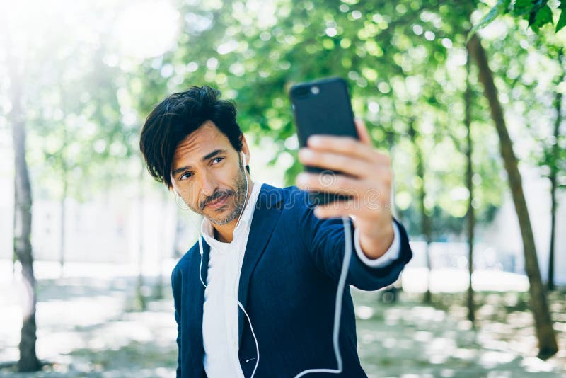 Привлекательный усмехаясь бизнесмен используя smartphone для listining музыки пока идущ в парк города Молодой человек делая selfi стоковое изображение Привлекательный усмехаясь бизнесмен используя smartphone для listining музыки пока идущ в парк города Молодой человек делая selfi стоковое изображение