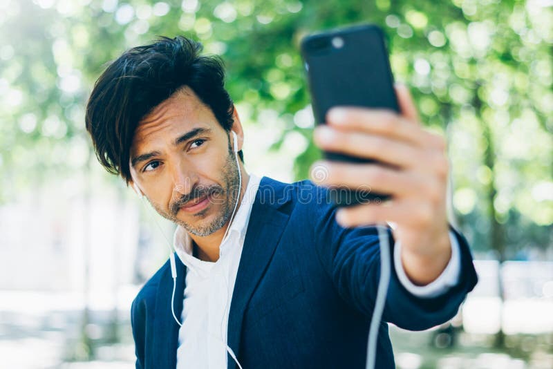 Привлекательный усмехаясь бизнесмен используя smartphone для listining музыки пока идущ в парк города Молодой человек делая selfi стоковое изображение rf Привлекательный усмехаясь бизнесмен используя smartphone для listining музыки пока идущ в парк города Молодой человек делая selfi стоковое изображение rf