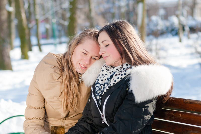 Two happy attractive charming girls sitting on a bench in winter where one of them leaned on the shoulder of another portrait. Two happy attractive charming girls sitting on a bench in winter where one of them leaned on the shoulder of another portrait