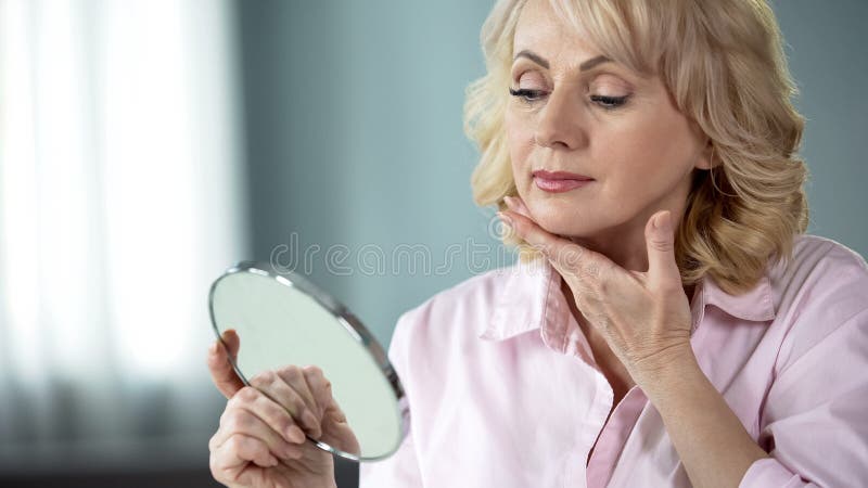Attractive aged woman looking at her face in mirror, plastic surgery effect, stock photo. Attractive aged woman looking at her face in mirror, plastic surgery effect, stock photo