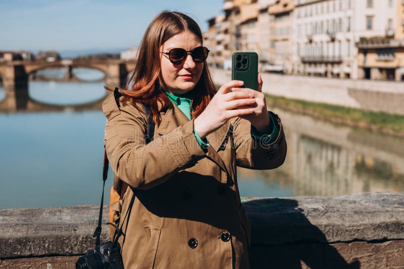 Attractive young female tourist is exploring new city. Redhead girl holding a smartphone on famous Old bridge in Florence. Traveling. Happy girl walking in city and makes selfie. Attractive young female tourist is exploring new city. Redhead girl holding a smartphone on famous Old bridge in Florence. Traveling. Happy girl walking in city and makes selfie