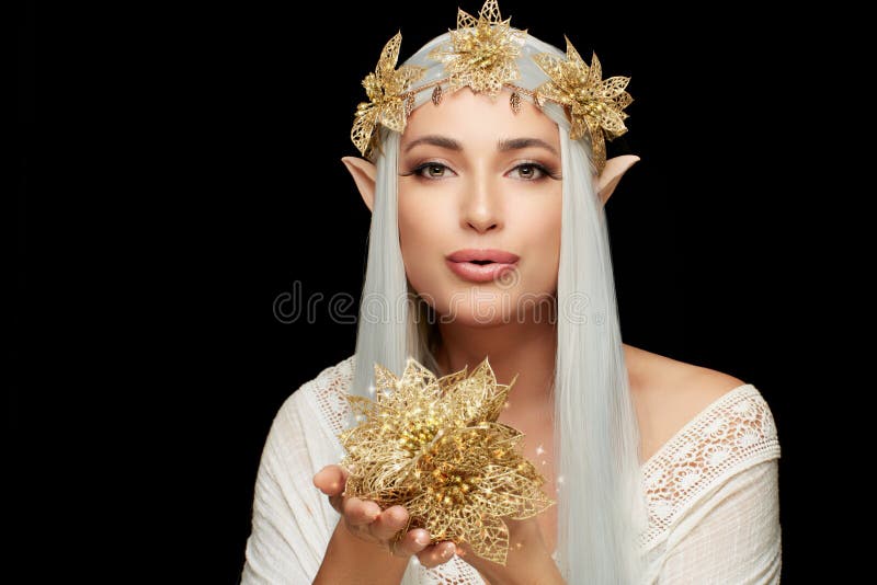Attractive young woman in elf queen costume with golden christmas poinsettia flower wreath looking at camera holding golden flowers on hans. Beautiful young woman as christmas fairy isolated on black. Attractive young woman in elf queen costume with golden christmas poinsettia flower wreath looking at camera holding golden flowers on hans. Beautiful young woman as christmas fairy isolated on black.