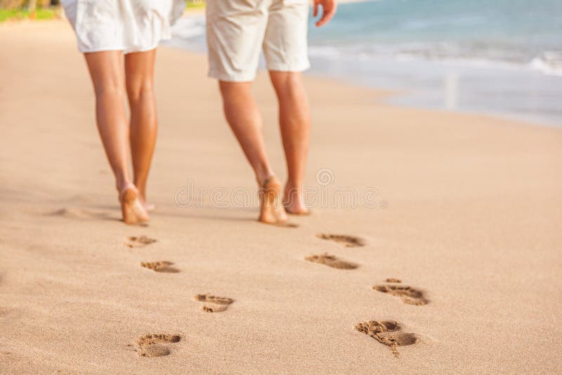 Beach couple relaxing at sunset walking barefoot. Focus on footprints in golden sand. Closeup of legs. Romantic beach vacation holidays. Young people from behind walking away towards happiness. Beach couple relaxing at sunset walking barefoot. Focus on footprints in golden sand. Closeup of legs. Romantic beach vacation holidays. Young people from behind walking away towards happiness.