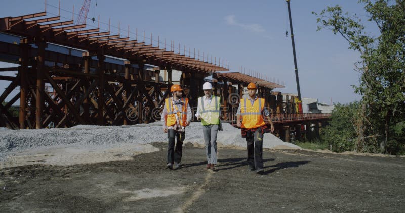 Tracking shot of mature and adult men in hardhats and waistcoats walking towards unfinished bridge and speaking with each other on sunny day. Tracking shot of mature and adult men in hardhats and waistcoats walking towards unfinished bridge and speaking with each other on sunny day