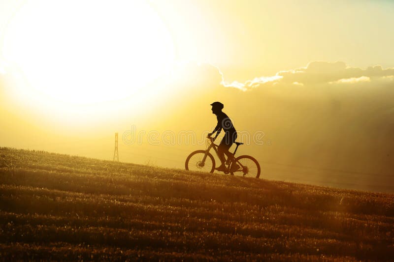 Profile silhouette of sport man cycling uphill riding cross country mountain bike on sunset field with harsh sun light and high contrast in amazing beautiful rural landscape with orange lensflare. Profile silhouette of sport man cycling uphill riding cross country mountain bike on sunset field with harsh sun light and high contrast in amazing beautiful rural landscape with orange lensflare