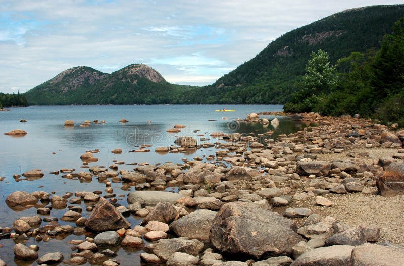 A low view of Jordan Pond in Maine with beige rocky shoreline in foreground and forested hills in background. A low view of Jordan Pond in Maine with beige rocky shoreline in foreground and forested hills in background.