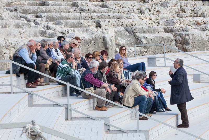 Travel guide with group of tourists sitting on ancient steps. A tourist guide visitors tourists is explaining the history of the greek theater of Syracuse in Sicily. Travel guide with group of tourists sitting on ancient steps. A tourist guide visitors tourists is explaining the history of the greek theater of Syracuse in Sicily.
