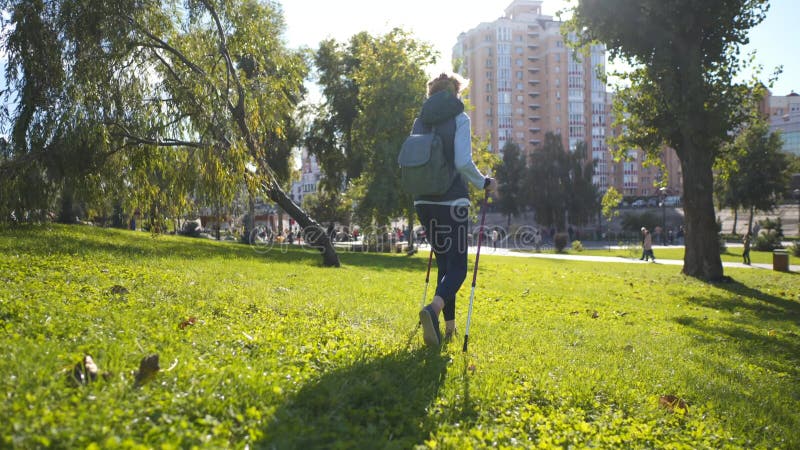 работоспособный и активный. зрелая женщина с прогулочными шестами прогуливается на лужайке в городском парке в солнечную погоду. в видеоматериал