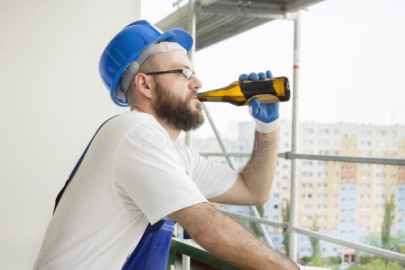Рабочий-строитель в носке работы, защитных перчатках и шлеме на голове выпивает пиво от бутылки Работа на большой возвышенности стоковое фото Рабочий-строитель в носке работы, защитных перчатках и шлеме на голове выпивает пиво от бутылки Работа на большой возвышенности стоковое фото