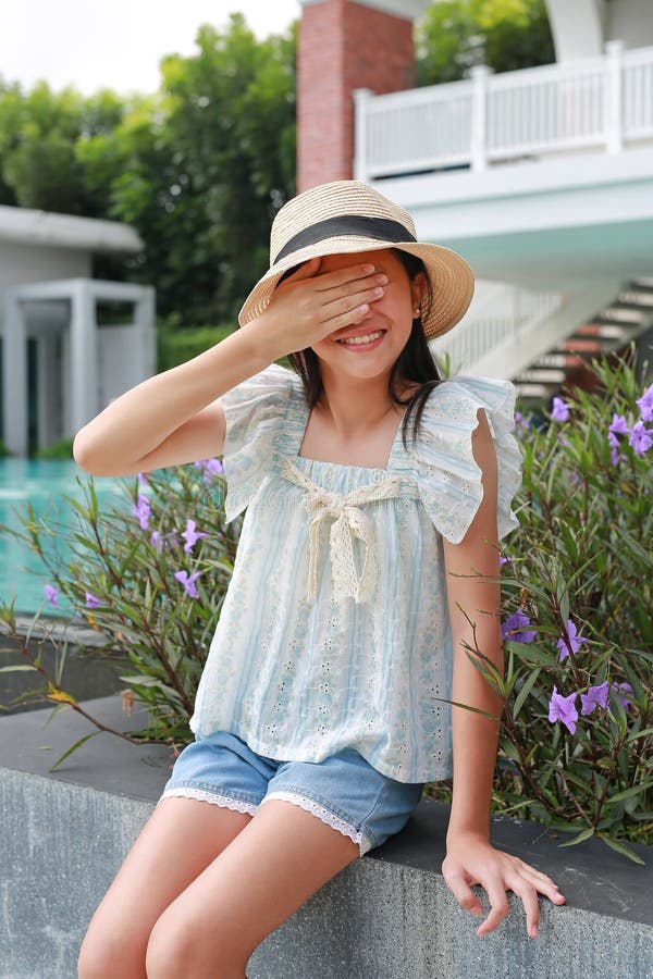 Радостная юная девочка в соломенной шляпе и летнем топе сидит у бассейна, закрывая глаза рукой в игривый, застенчивый или солнечны стоковая фотография rf