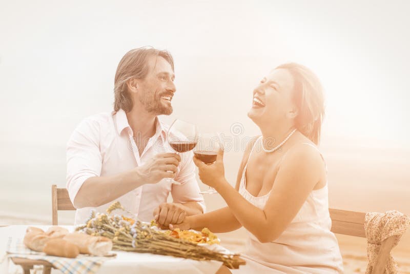 Радостное вино напитка, мужчины и женщины в кафе на пляже. Прекрасные пары смеясь держаться за руки и наслаждаясь жизнью, сидя  стоковая фотография