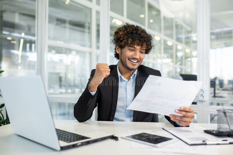 A young, curly-haired businessman exudes joy while looking at documents in a modern office setting, making a triumphant gesture. A young, curly-haired businessman exudes joy while looking at documents in a modern office setting, making a triumphant gesture.