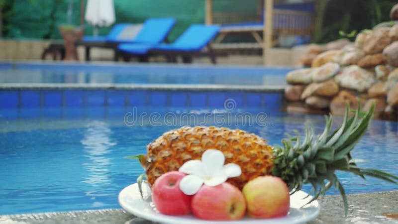 Различные плодоовощи ананас, яблоки, красивый цветок frangipani положены на плиту на задней части бассейна