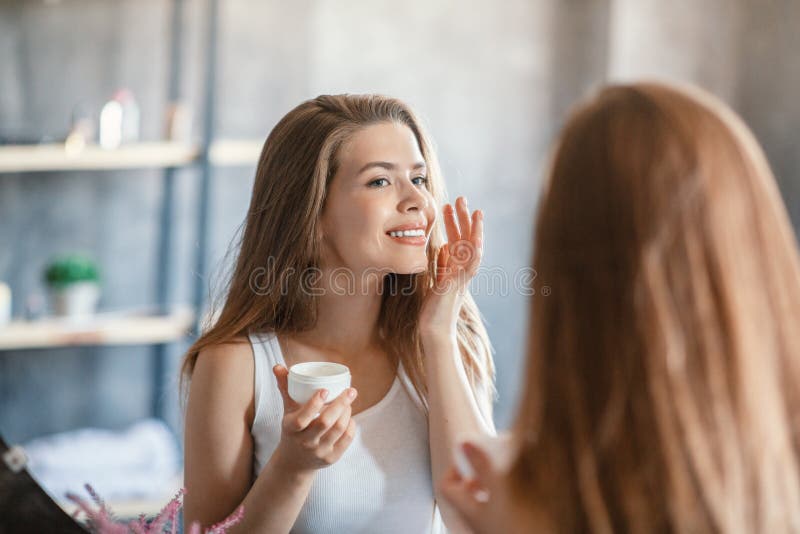 Режим по уходу за кожей. Прекрасная белокурая девочка, применяющим лицевую сливки около зеркала в ванной стоковое фото rf