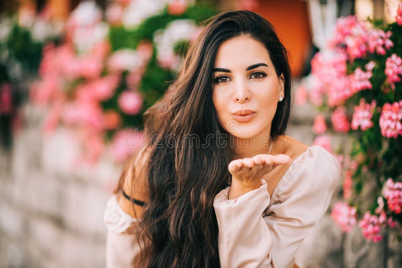 Close up portrait of beautiful young woman with long shiny hair, posing outdoor in pink flowers, sending a kiss. Close up portrait of beautiful young woman with long shiny hair, posing outdoor in pink flowers, sending a kiss