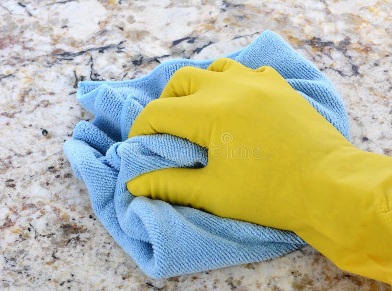 Closeup of a hand in a yellow latex glove using a blue towel to clean a granite counter top. Closeup of a hand in a yellow latex glove using a blue towel to clean a granite counter top.