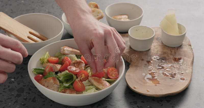 Рука человека добавляет помидоры черри и курицу в салат цезаря в белом керамическом шаре на конкретном countertop стоковые изображения rf