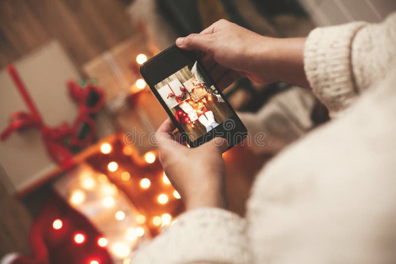 Руки держа телефон и принимая фото подарочных коробок рождества, шляпу santa, света на деревянной предпосылке в темной комнате Ст стоковые изображения rf Руки держа телефон и принимая фото подарочных коробок рождества, шляпу santa, света на деревянной предпосылке в темной комнате Ст стоковые изображения rf