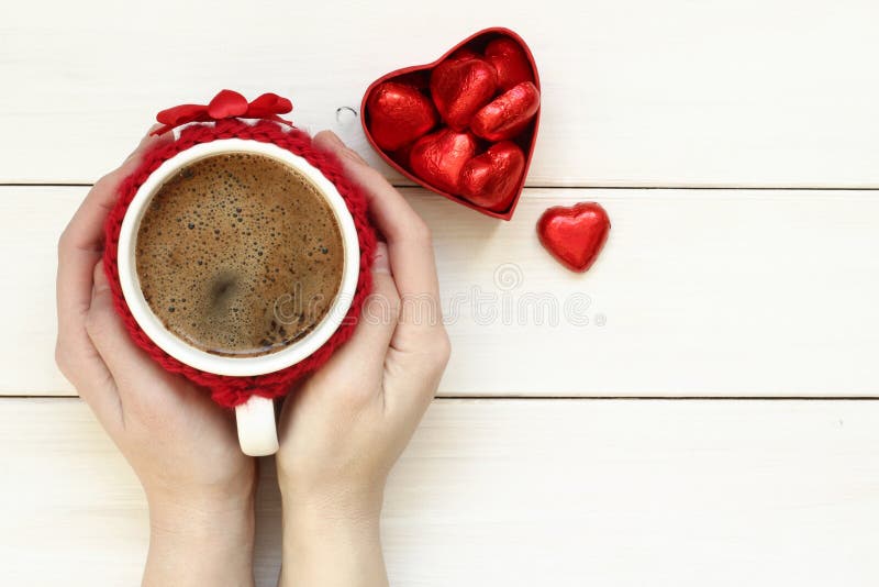Woman hands holding cup of hot coffee with red knitted warmer with bow and heart. Box of chocolate hearts on white wooden table. Valentines Day background. Woman hands holding cup of hot coffee with red knitted warmer with bow and heart. Box of chocolate hearts on white wooden table. Valentines Day background.