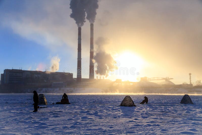 Рыбная ловля зимы, страсть людей, рыболовы улавливает рыб на замороженном реке на фоне труб фабрики на заходе солнца стоковые фотографии rf Рыбная ловля зимы, страсть людей, рыболовы улавливает рыб на замороженном реке на фоне труб фабрики на заходе солнца стоковые фотографии rf