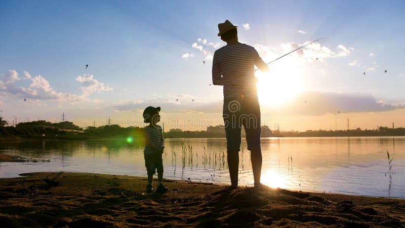 Father and son fishing on the riverside in the rays of setting sun - The child is smiling - Birds flying in the sky - Mid shot. Father and son fishing on the riverside in the rays of setting sun - The child is smiling - Birds flying in the sky - Mid shot