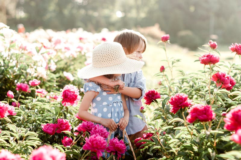 Сute children boy and girl walk among the flowers of blossoming peonies.