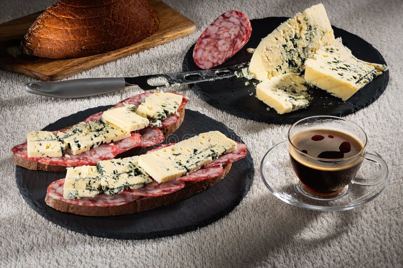 Сандвичи с сыром Dorblu и сырцовой копченой сосиской на черных плитах шифера, ноже сыра и чашке горячего кофе стоковое изображение rf Сандвичи с сыром Dorblu и сырцовой копченой сосиской на черных плитах шифера, ноже сыра и чашке горячего кофе стоковое изображение rf