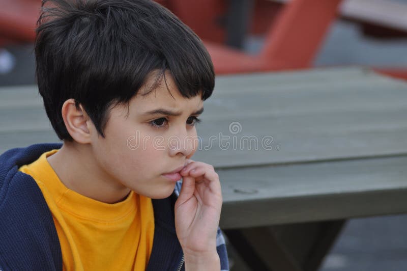 Little boy biting nails, worrying, while watching a group of kids play without him. He could be excluded from the group or in time out. Little boy biting nails, worrying, while watching a group of kids play without him. He could be excluded from the group or in time out.