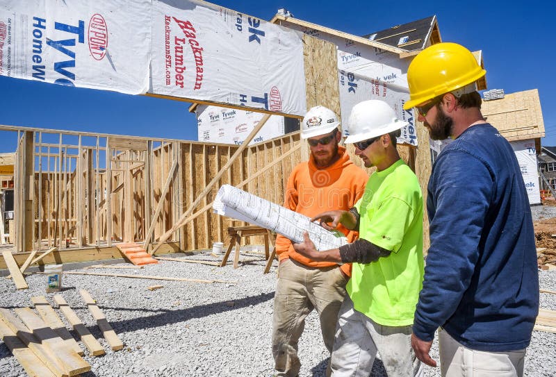 Three male construction workers, carpenters, wearing hardhats look at blueprints at new residential subdivision home building work site. Three male construction workers, carpenters, wearing hardhats look at blueprints at new residential subdivision home building work site