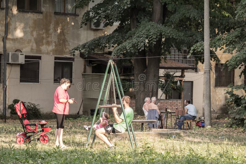 BELGRADE, SERBIA - AUGUST 2, 2015:  ..Picture of the children playground of the residential area of Staro Sajmiste, in belgrade, Serbia, with a Serbian family playing while old men are sitting behind. BELGRADE, SERBIA - AUGUST 2, 2015:  ..Picture of the children playground of the residential area of Staro Sajmiste, in belgrade, Serbia, with a Serbian family playing while old men are sitting behind