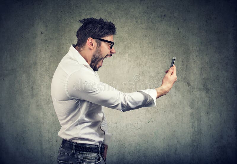 Side view of angry man holding smartphone and shouting in anger on gray background. Side view of angry man holding smartphone and shouting in anger on gray background.