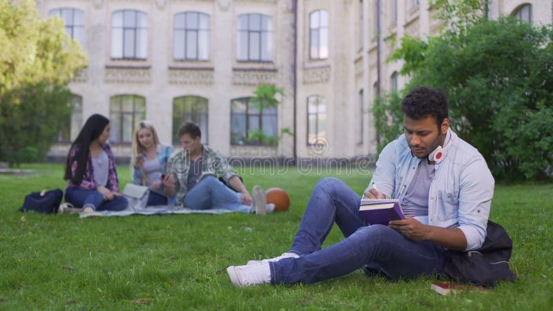 Серьезный biracial студент сидя на траве и писать эссе, делая домашнюю работу