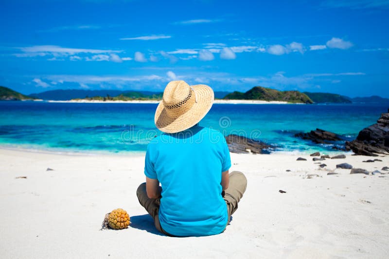 Man with hat sitting on the white sand tropical beach, looking at the coral island on the horizon. Man with hat sitting on the white sand tropical beach, looking at the coral island on the horizon