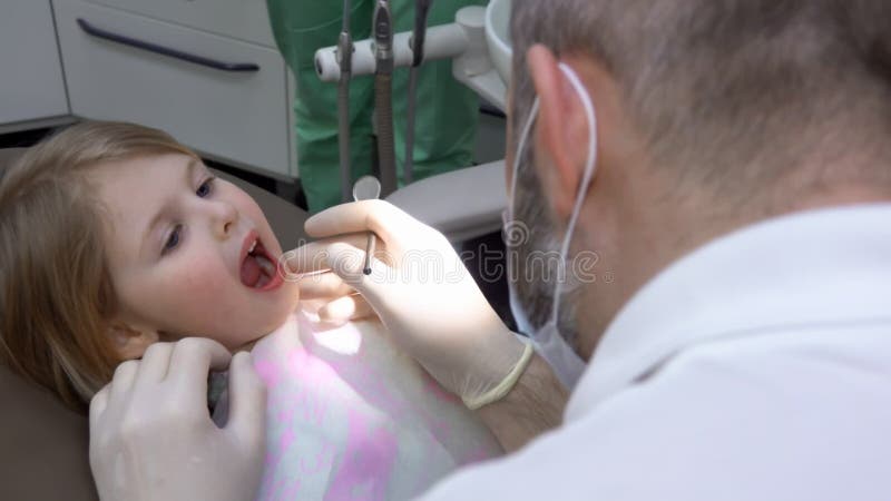 симпатичная маленькая девочка широко открывает рот, чтобы позволить зубному осмотреть зубы видеоматериал - Видео насчитывающей работа, гигиена: 232738574