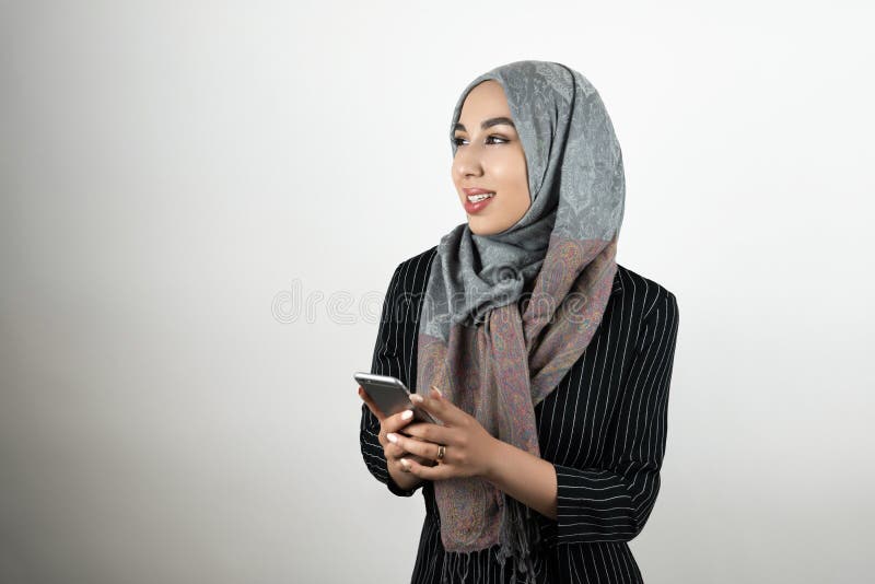 Смартфон удерживания головного платка hijab тюрбана молодой красивой мусульманской женщины нося в ее предпосылке изолированной ру стоковые фотографии rf Смартфон удерживания головного платка hijab тюрбана молодой красивой мусульманской женщины нося в ее предпосылке изолированной ру стоковые фотографии rf