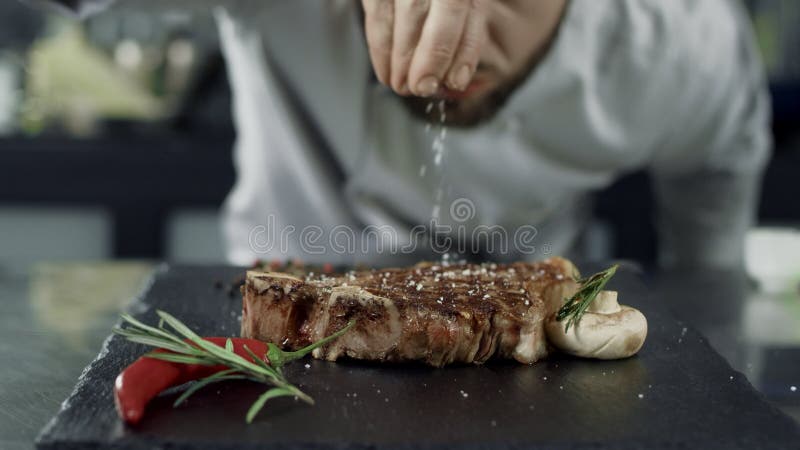 Соль шеф-повара жаря мясо на griddle Стейк соли рук человека крупного плана в замедленном движении