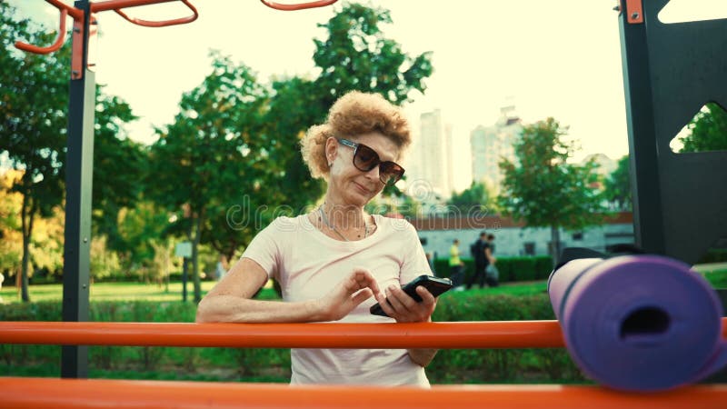 Спортивная зрелая женщина пользуется телефоном в уличном спортзале после занятий йогой. старшая кавказская леди отдыхает после зан видеоматериал