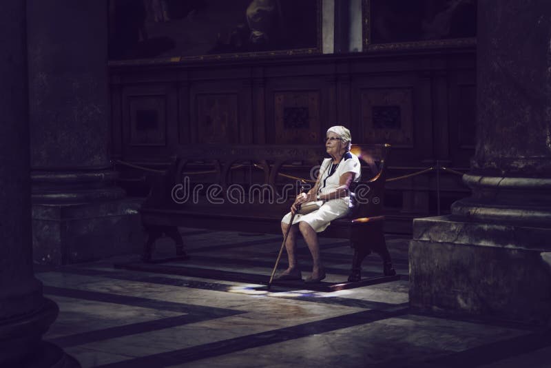 Old woman with a cane sitting on a church bench in the cathedral of Pisa, Italy. Colored light from a window in the ceiling enlightens her. Old woman with a cane sitting on a church bench in the cathedral of Pisa, Italy. Colored light from a window in the ceiling enlightens her.