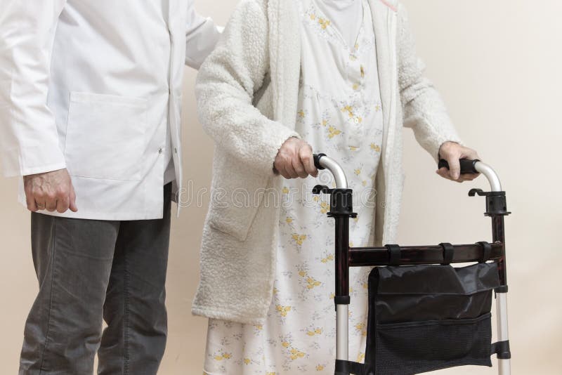 A senior women in a white bathrobe and nightgown goes with a rehabilitation walker. The nurse supports and protects the old woman. A senior women in a white bathrobe and nightgown goes with a rehabilitation walker. The nurse supports and protects the old woman.