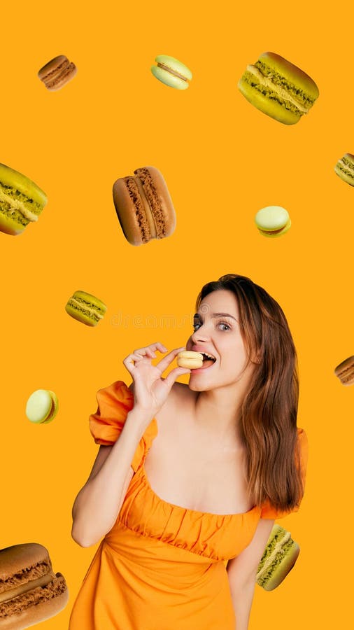 Стильная молодая женщина в оранжевом платье, кусающая макарон, окруженная плавающими разноцветными лакомствами на оранжевом фоне. стоковая фотография