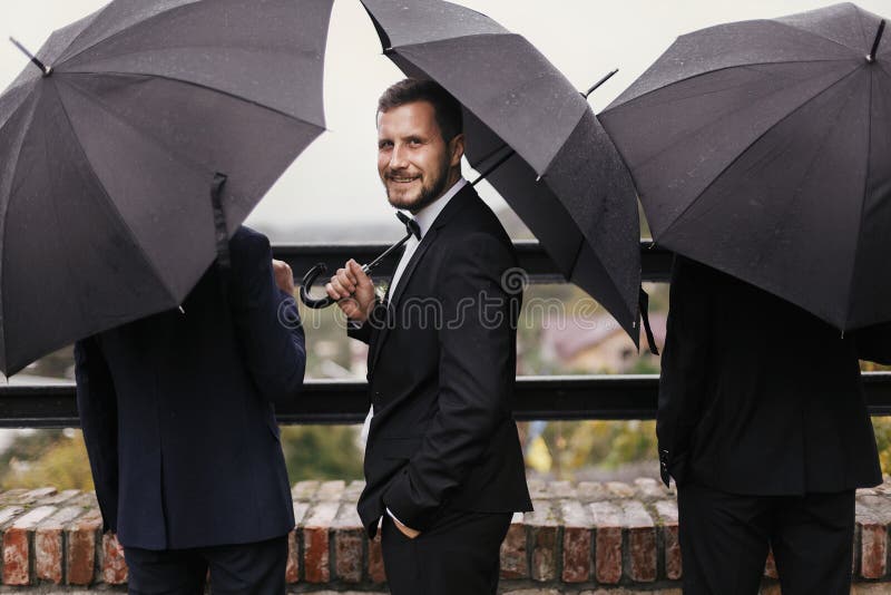Стильный выхольте и groomsmen стоя под черным зонтиком и po стоковая фотография Стильный выхольте и groomsmen стоя под черным зонтиком и po стоковая фотография