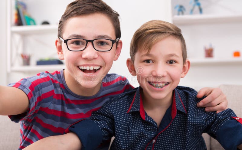 Two handsome teenage boys taking selfie while posing with smile to phone at home. Wearing glasses and braces. Friendship, brotherhood and health care concept. Two handsome teenage boys taking selfie while posing with smile to phone at home. Wearing glasses and braces. Friendship, brotherhood and health care concept