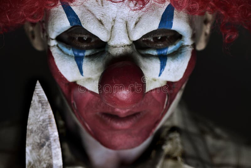 Closeup of a scary evil clown wearing a ragged shirt, holding a big knife next to his face. Closeup of a scary evil clown wearing a ragged shirt, holding a big knife next to his face