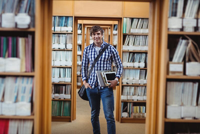 Студент стоя в библиотеке с компьтер-книжкой и цифровой таблеткой стоковая фотография