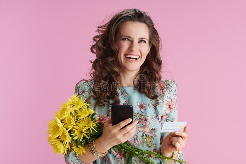 Happy modern middle aged woman with long wavy brunette hair with yellow chrysanthemums flowers and credit card sending text message using smartphone isolated on pink. Happy modern middle aged woman with long wavy brunette hair with yellow chrysanthemums flowers and credit card sending text message using smartphone isolated on pink