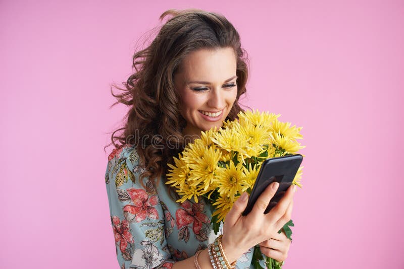 happy modern woman in floral dress with yellow chrysanthemums flowers sending text message using smartphone against pink background. happy modern woman in floral dress with yellow chrysanthemums flowers sending text message using smartphone against pink background