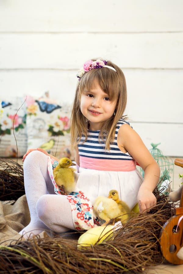 Счастливая маленькая девочка в платье с цветками на ее голове сидит в гнезде и держит милых пушистых утят пасхи на ее коленях