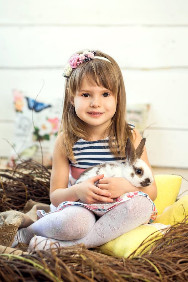 Счастливая маленькая девочка в платье сидит в гнезде и держит милый пушистый белый зайчика пасхи