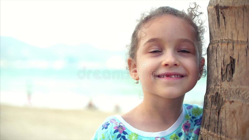 Счастливая маленькая девочка на пляже в голубом платье с пальмой, смотрящ камеру, усмехаться и немного застенчивое Ребенок