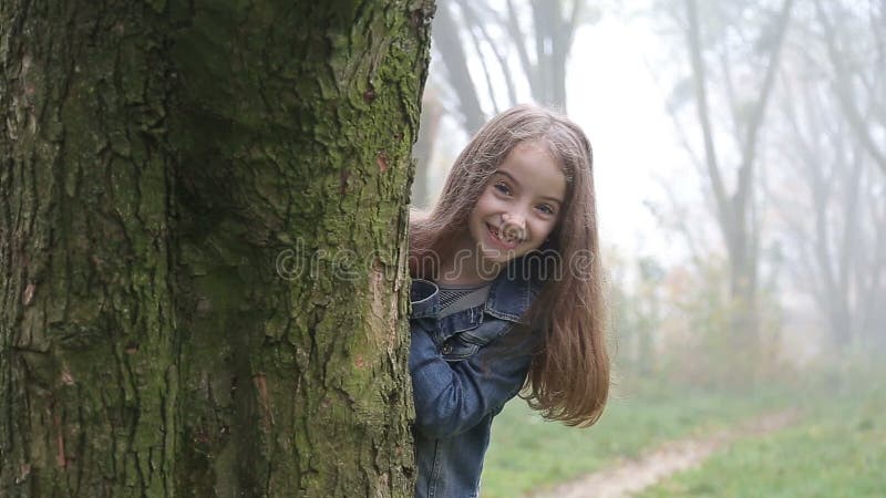 Счастливая маленькая девочка смотрит от заднего дерева и улыбок к камере